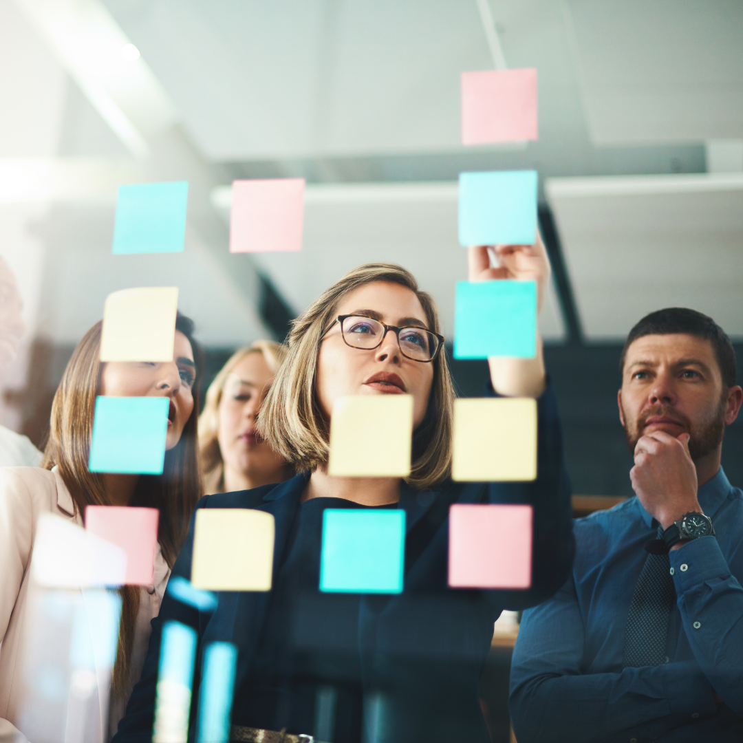 A team of professionals stands behind a glass wall covered in colourful sticky notes. A woman in glasses leads a planning session, pointing at the notes while the team watches attentively.